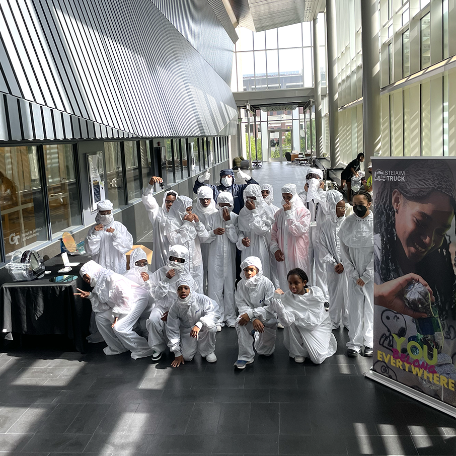 A group of Georgia Tech students dressed in cleanroom protective gear wait to greet secondary students as they arrive for a tour of facilities and research labs.